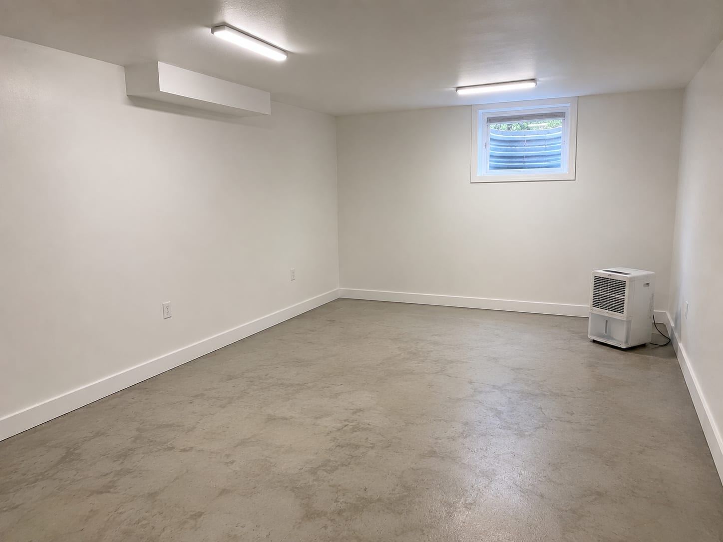 Freshly finished and painted basement room with new drywall, clean concrete floor, and a dehumidifier running in the corner after completed remediation