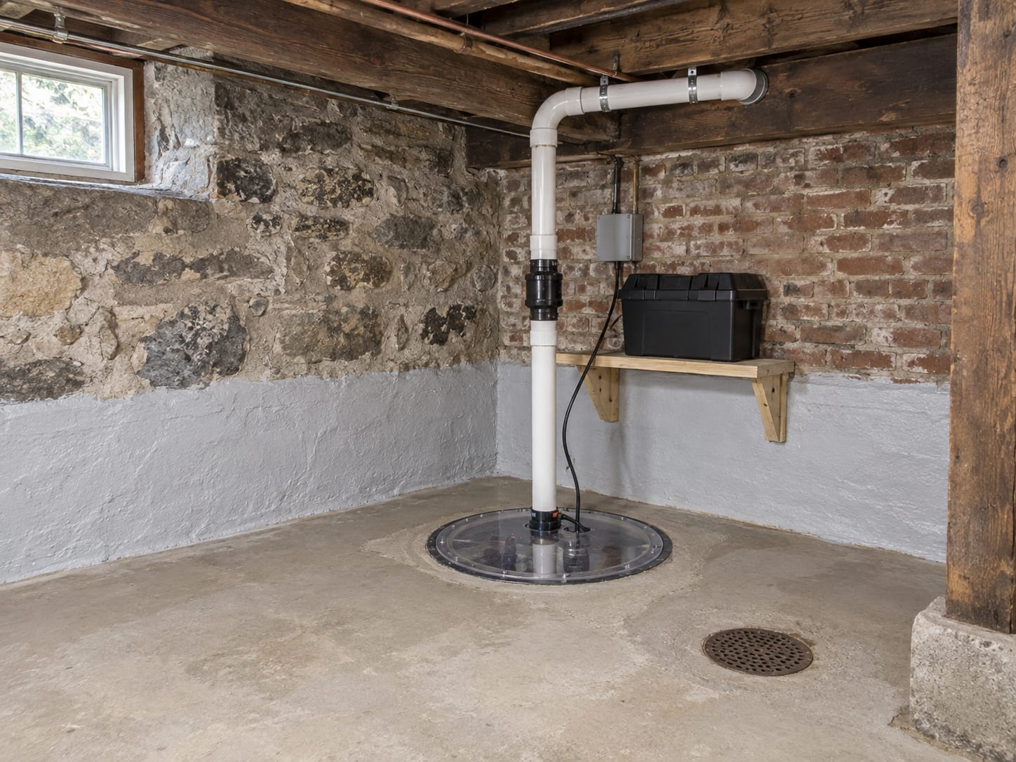 A sealed sump pit with a lid, a battery backup unit and a PVC discharge pipe through the rim joist, in the brick foundation cellar of a Rocky Hill, New Jersey house