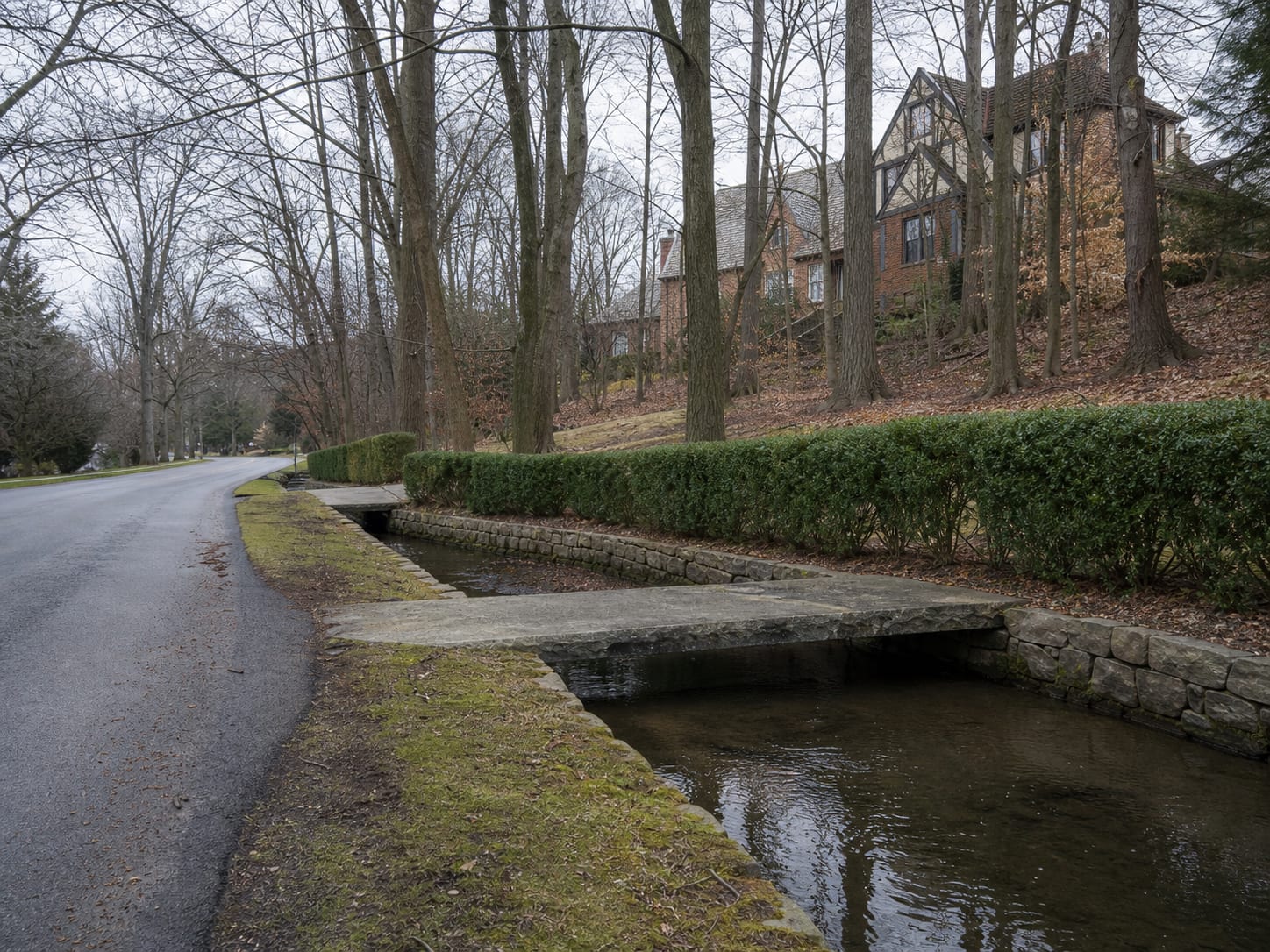 A quiet residential road in Short Hills, New Jersey running beside a stone walled brook channel, with clipped hedges, deep setbacks and slate roofed houses set back among mature trees