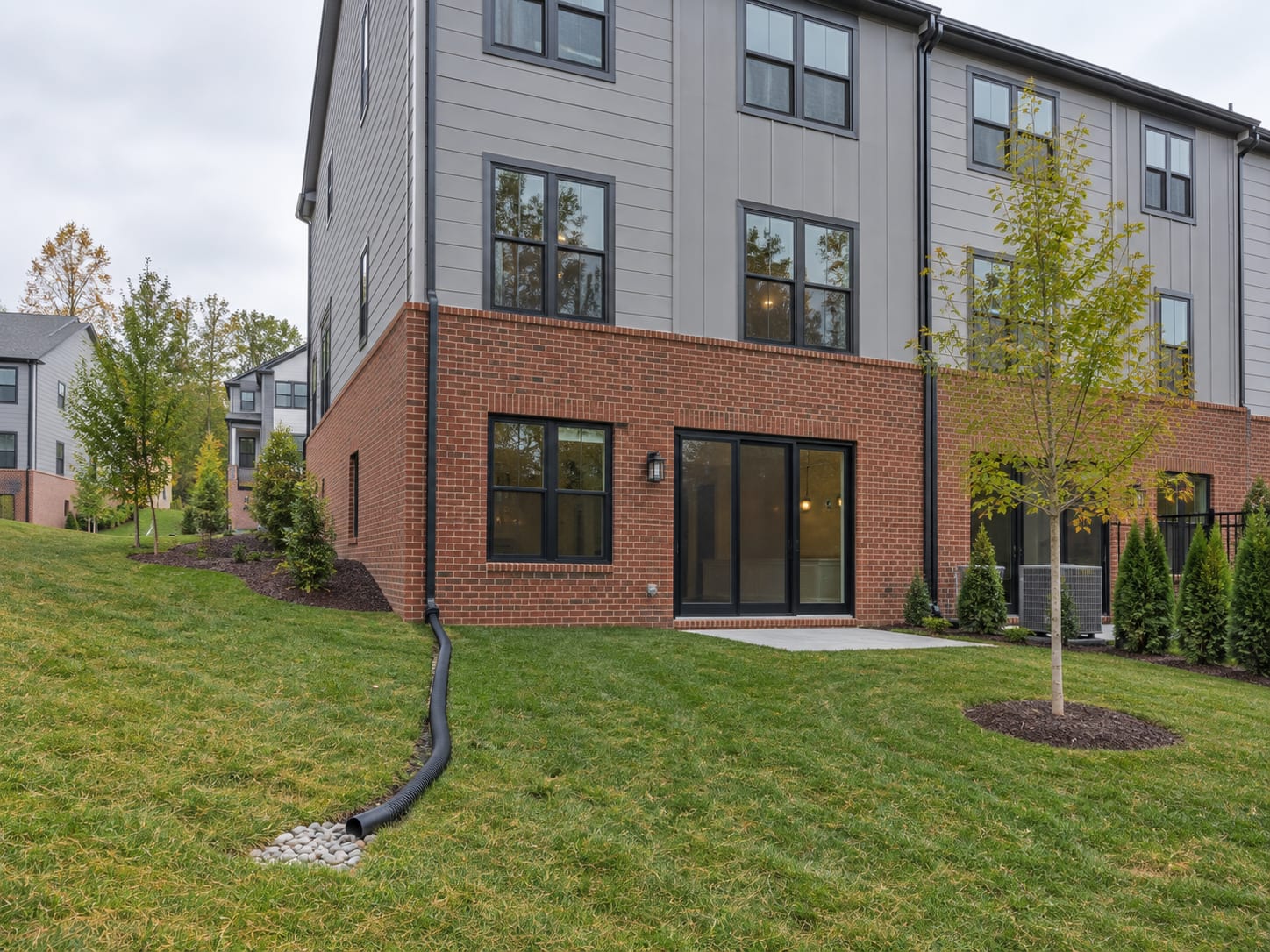 The rear of a brick and fiber cement townhouse with a gutter, a downspout and a buried extension pipe discharging onto sloped grass, in Skillman, New Jersey
