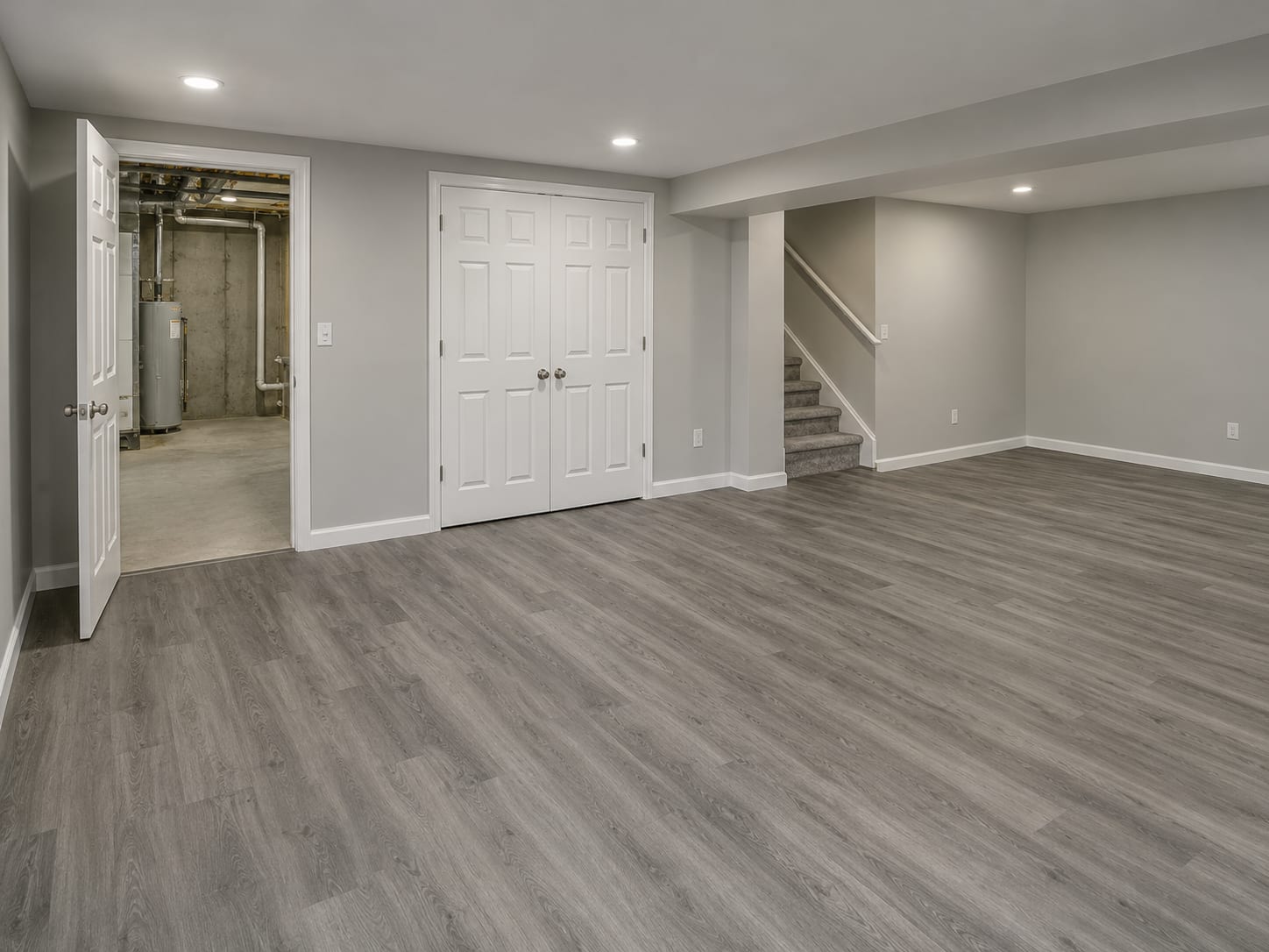 An empty finished basement room with new primed drywall, vinyl plank flooring and recessed lights, a closet door and a stair, in Ten Mile Run, New Jersey