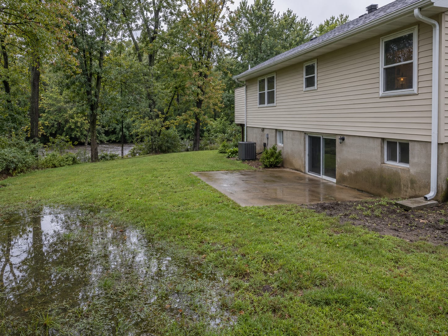 A back lawn behind a raised ranch holding standing water in the low corner with a wooded stream line behind and a downspout at the house, in Ten Mile Run, New Jersey