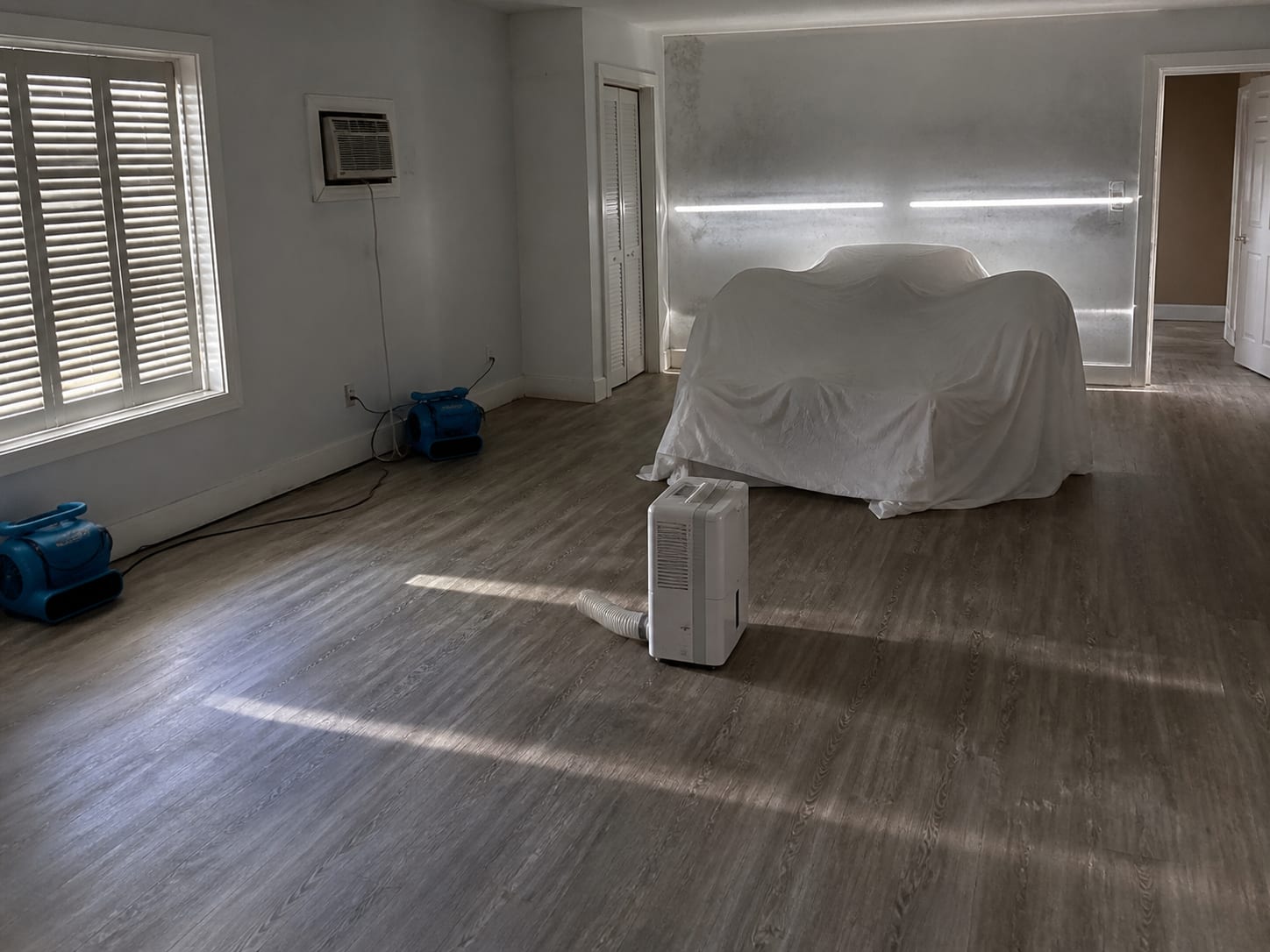 A dehumidifier and air movers running in a shuttered ground floor seasonal rental unit in Wildwood, New Jersey, with slatted light coming through closed storm shutters