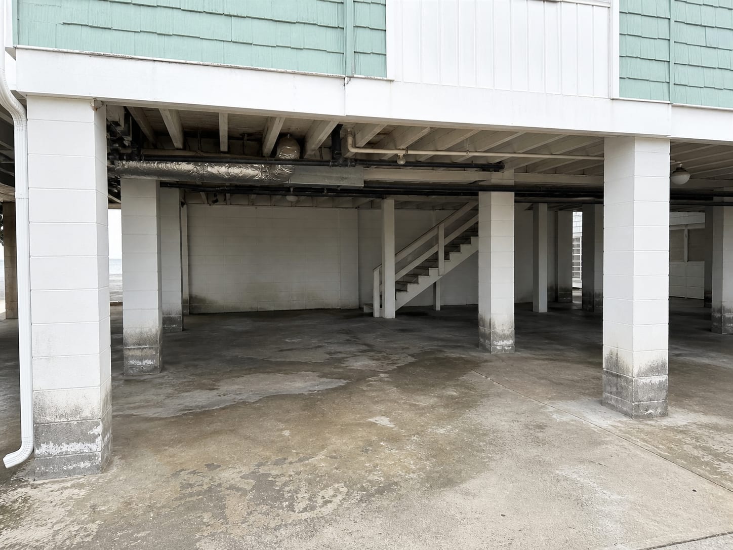 An open at-grade parking bay tucked under the living floors of a Wildwood, New Jersey building, with the underside of the floor above exposed to outside air and damp staining rising up the block piers