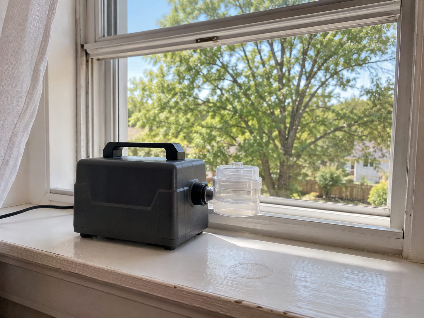 Spore trap cassette sampling pump resting on a clean wooden windowsill inside a New Jersey home next to an open window showing a mature deciduous tree outside