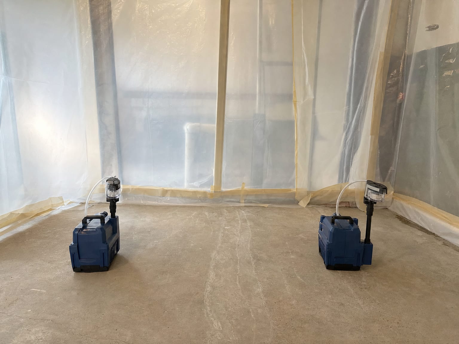 Two air sampling pumps with cassettes positioned on a clean concrete floor inside a sealed plastic containment barrier in a basement